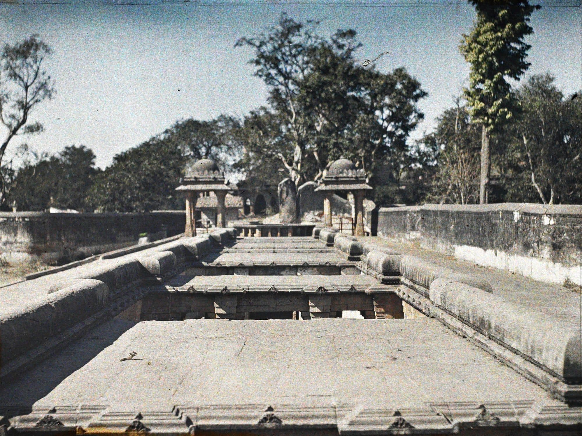 A colour autochrome looking along the top of a stepwell: flat stone roof slabs over a stepped corridor with openings down into it, low carved parapets on either side, two small domed kiosks at the far end and tall trees behind under a blue sky.