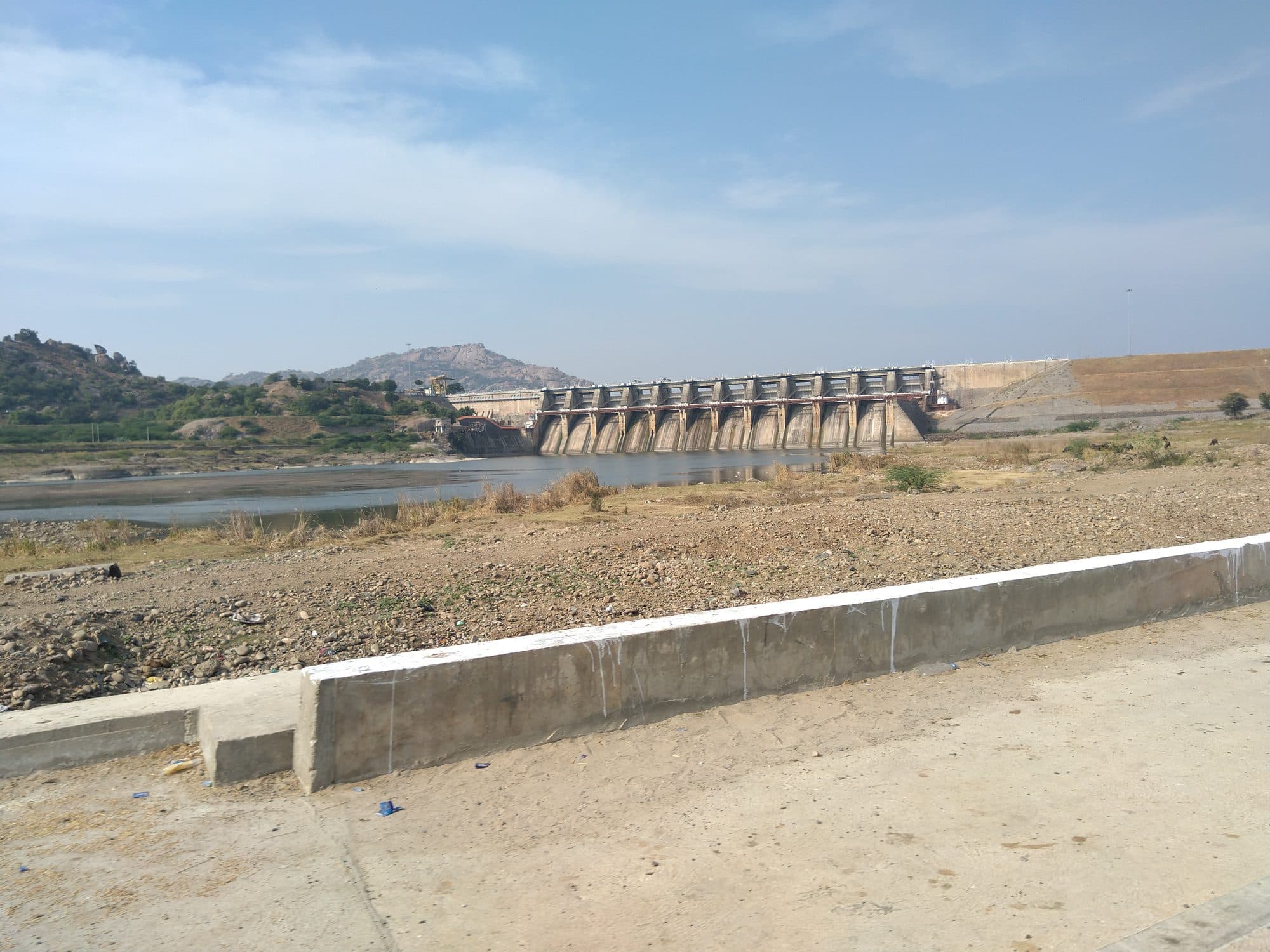 The gated concrete spillway of Dharoi dam seen across a stony, dry riverbed with a shallow pool of water at its foot, rocky hills to the left and an earthen embankment to the right, under a partly cloudy sky.