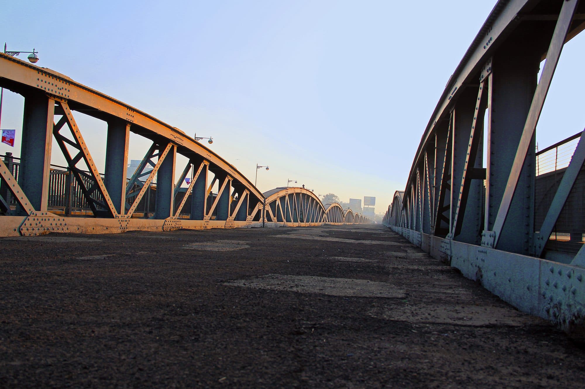 A low view along the deck of the Ellis Bridge: riveted steel bowstring trusses curve up on either side and repeat into the distance, with old lamp posts along the top chords and a hazy morning sky.