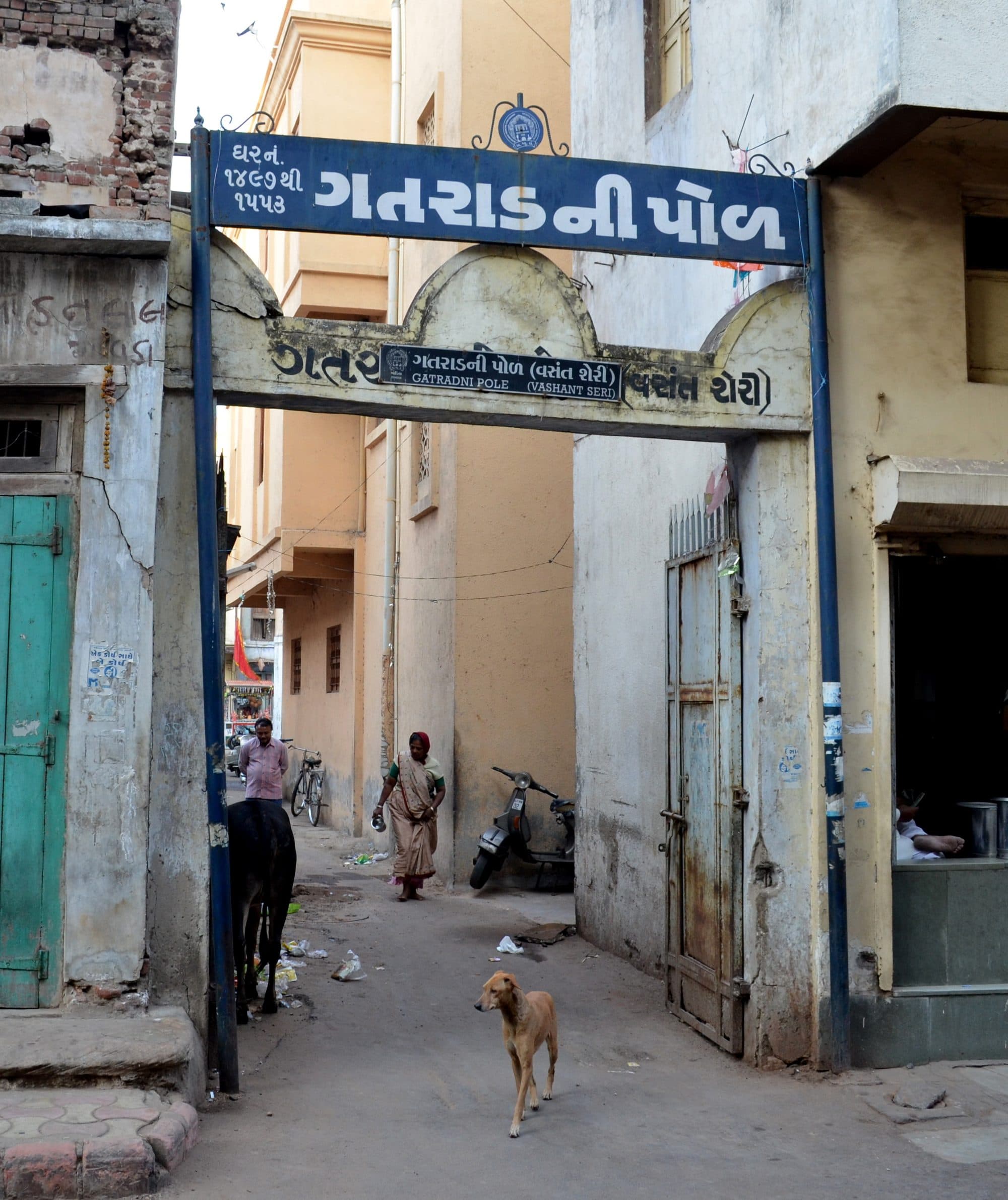 A pol gate in Ahmedabad: a painted blue signboard in Gujarati across the top of a plastered arched gateway, opening onto a narrow lane between tall attached houses. A woman carrying a small vessel walks towards the gate, a man stands by a bicycle, and a dog and a cow are in the foreground.