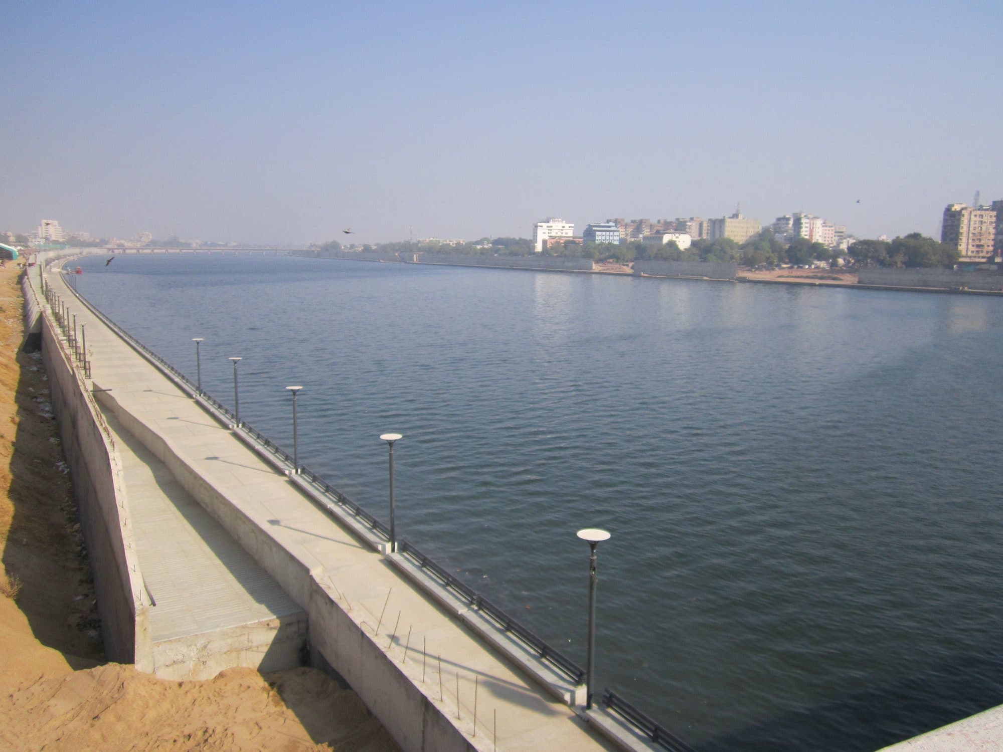 A broad, full river between straight concrete embankments. On the near bank a lower promenade with lamp posts runs along the water below a retaining wall and a strip of sand; buildings line the far bank and a long bridge crosses in the distance under a hazy blue sky.