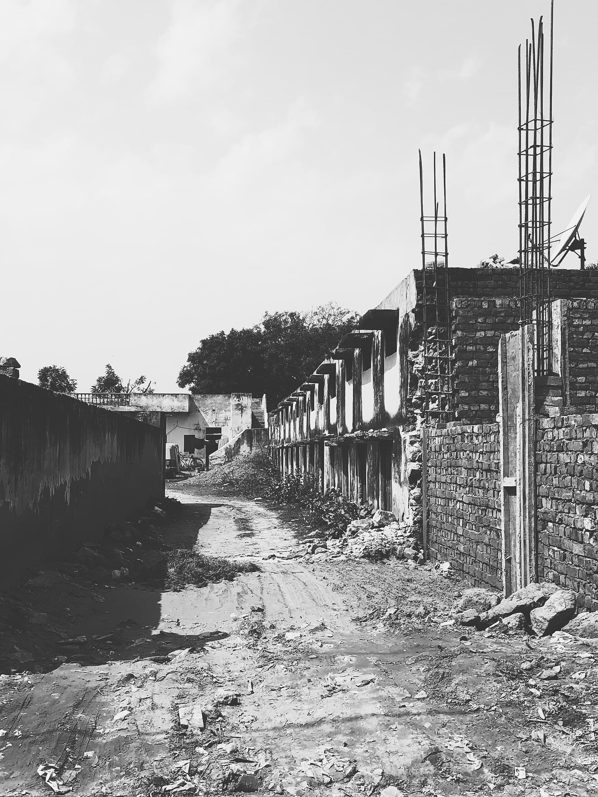 A black and white photograph of an unpaved, rutted lane in the village of Behrampur in Gurugram. On the right a terrace of half-built brick houses runs into the distance, unrendered, with reinforcing bars projecting from the unfinished roof and a satellite dish already mounted. Rubble and building debris line both sides of the track. A plastered compound wall closes the left side.