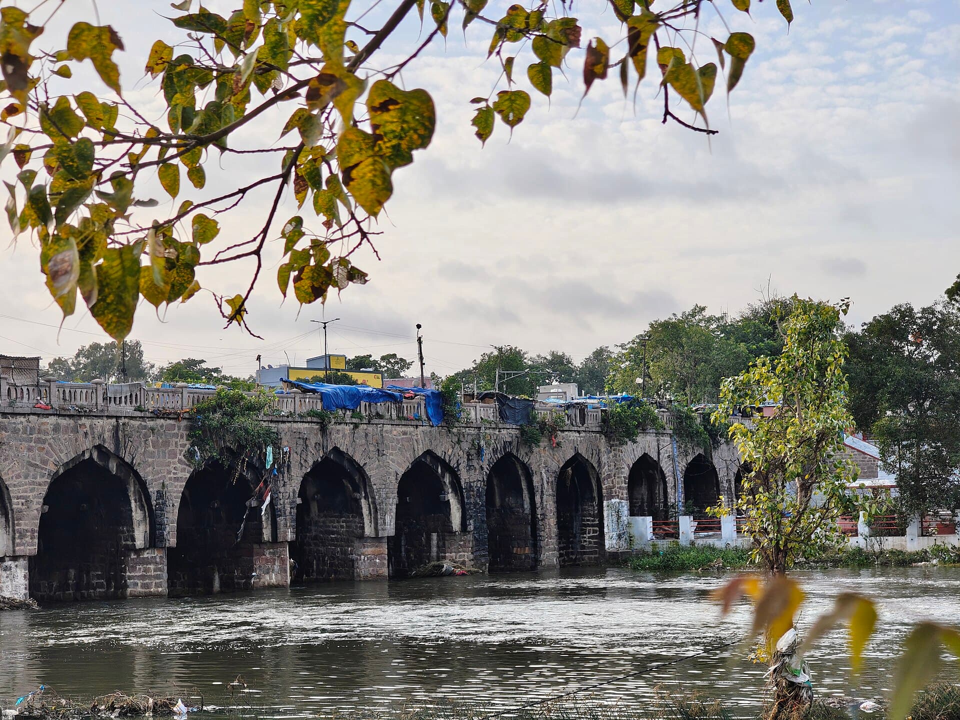 The Musi river at Purana Pul, Hyderabad. A long arched stone bridge crosses the frame with shelters and shops built along its parapet; the water below carries visible debris along its edge.