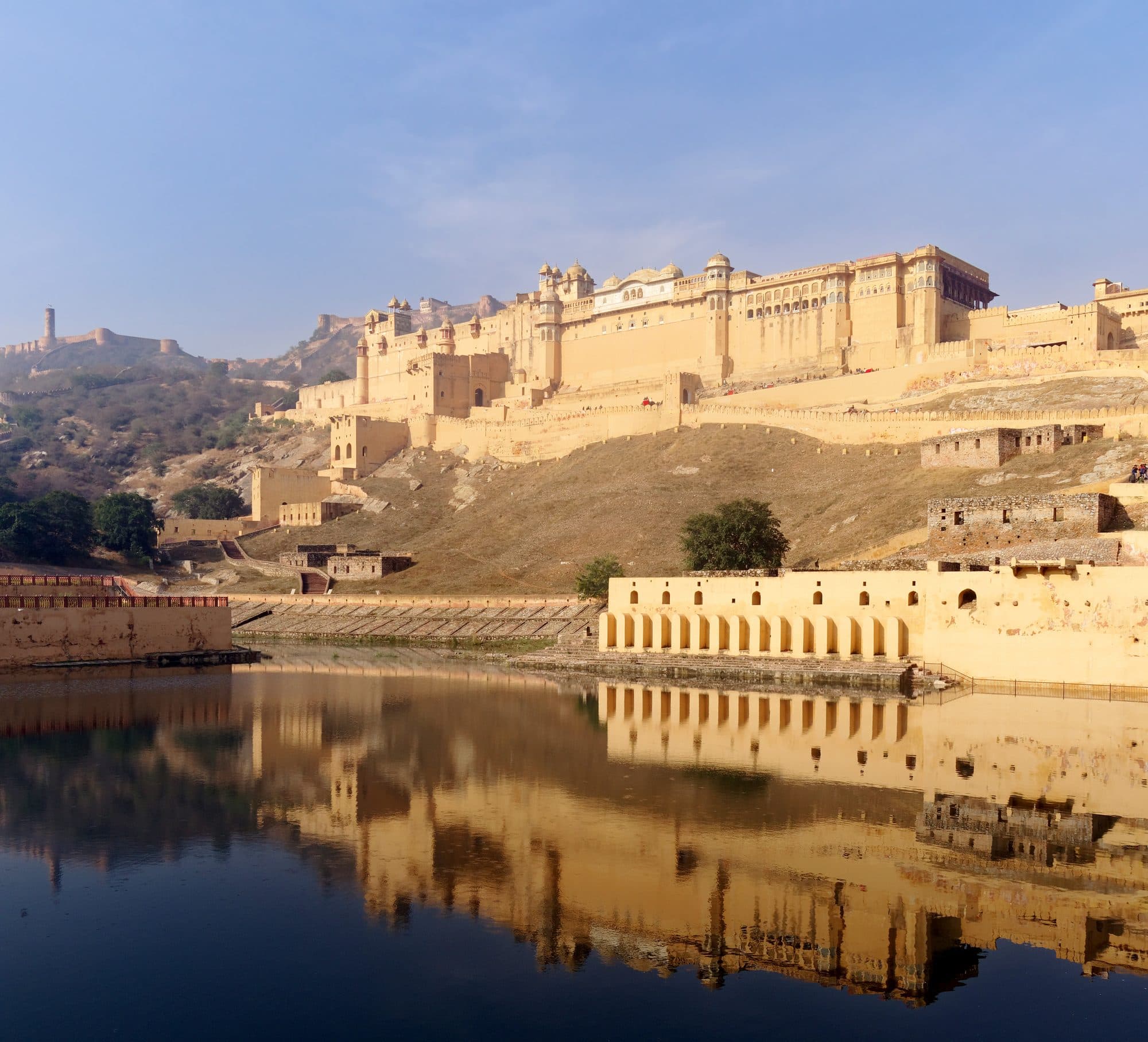 Amer Fort, a long sandstone palace on a hillside, reflected in the still blue water of Maota lake. An arcaded stone wall runs along the near shore below the fort. On the ridge far to the left, a second fort's walls and a watchtower stand against the sky.