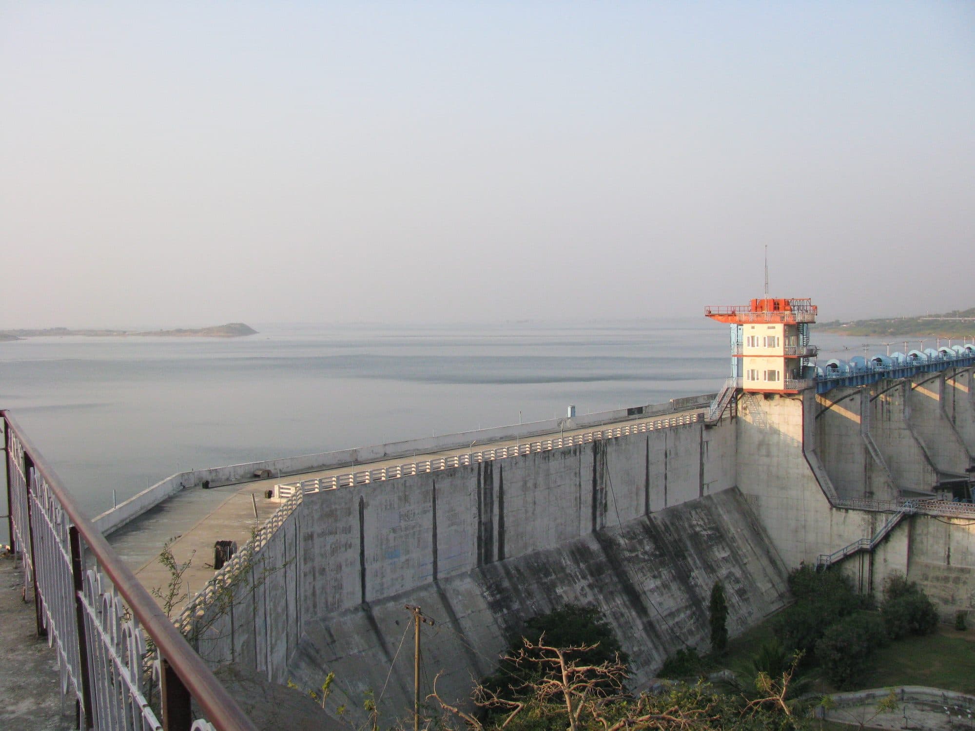 A tall concrete dam wall with a road along its crest and an orange-and-white control tower, holding back a wide, calm reservoir that stretches to a hazy horizon with a low island. A railing runs along the near edge.