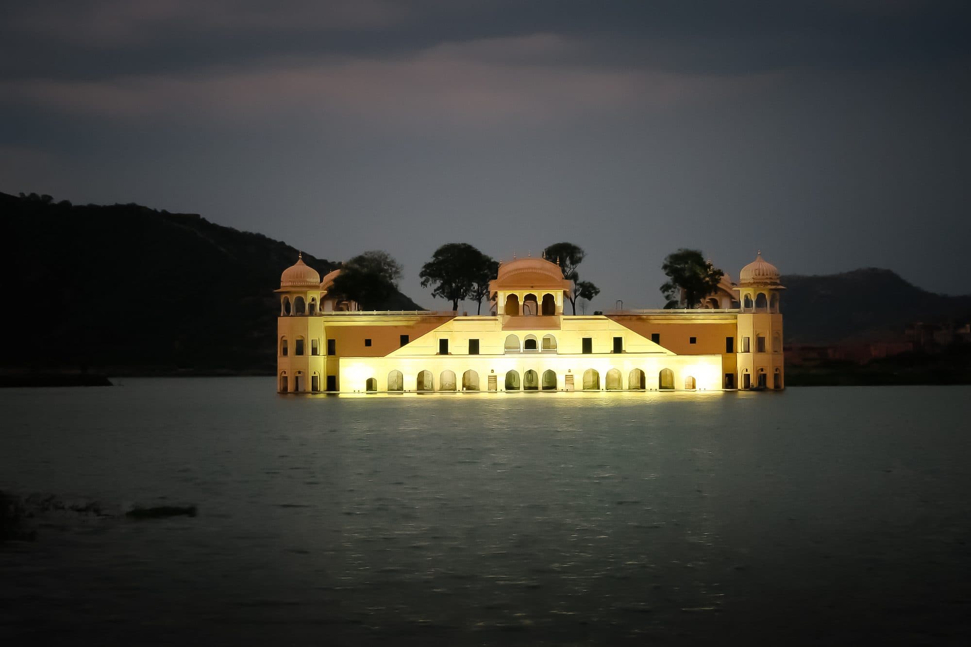 Jal Mahal lit up in warm light in the middle of Man Sagar lake, its reflection on the dark water, with black hills and a dark grey sky behind.