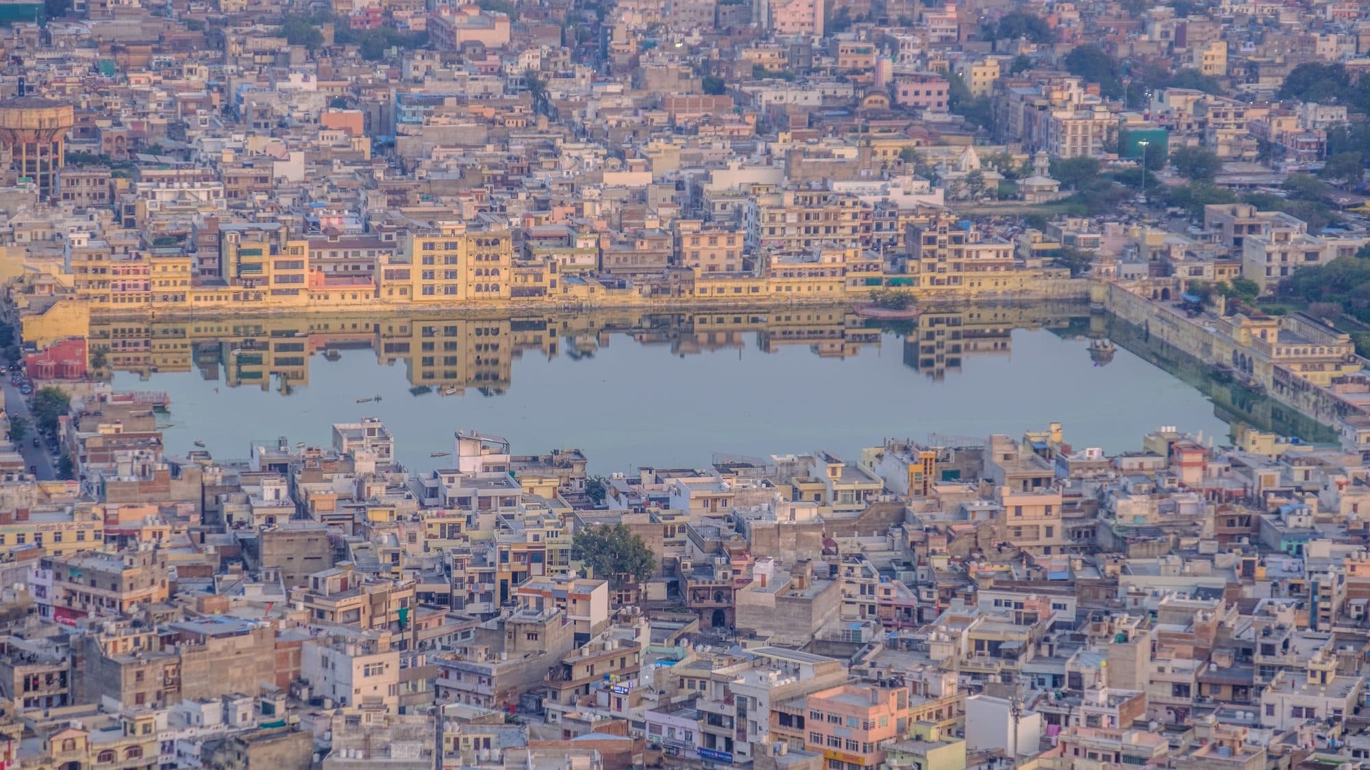 An aerial view over dense rows of cream and yellow city buildings. In the middle lies a long rectangular tank of still blue water with straight walled edges, the buildings around it reflected in the surface.