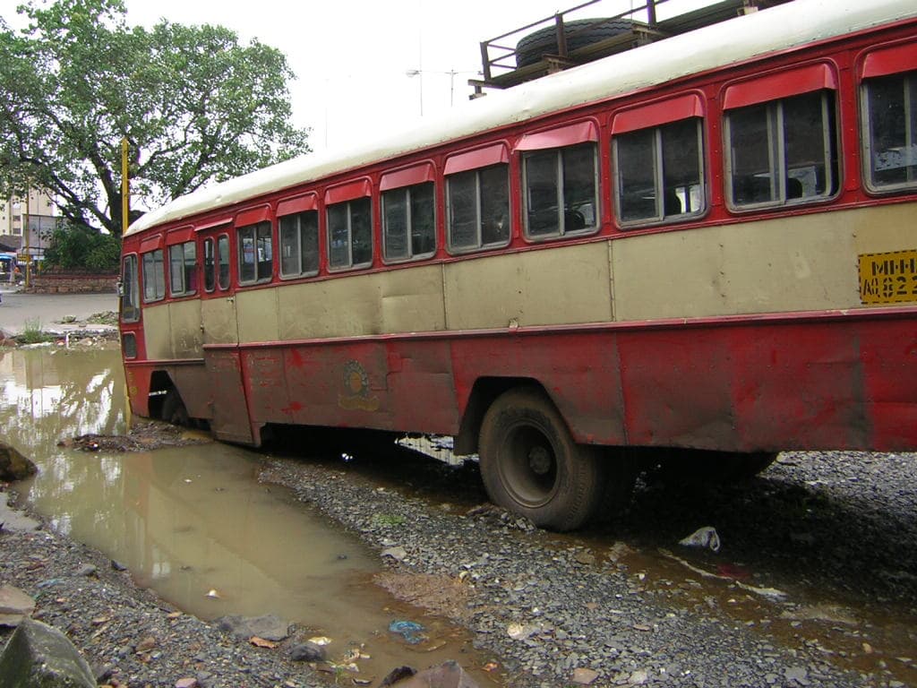 A red state-transport bus abandoned at an angle on a wrecked road near a flyover, debris and stranded vehicles around it.