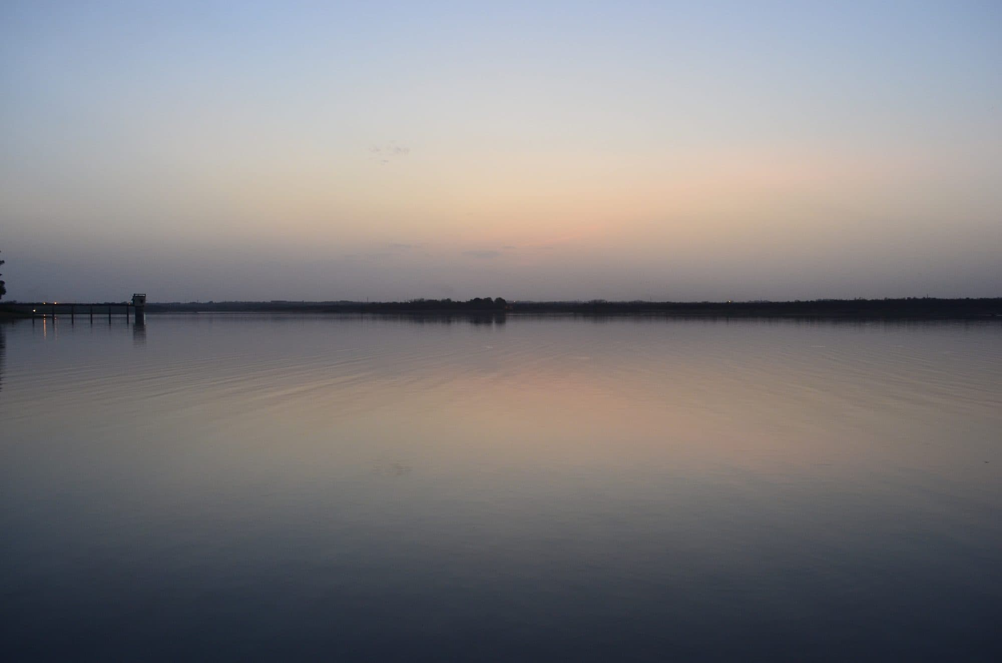 A wide, calm lake at dusk under a pale peach and blue sky, with a low line of trees on the far shore. At the far left edge a small tower stands in the water at the end of a walkway.