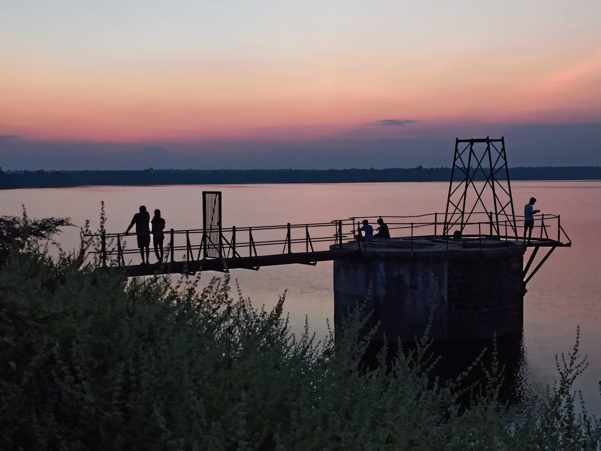 Silhouettes at dusk: a masonry tower stands in a calm lake reached by a railed metal walkway, with a steel frame on top. Several people stand on the walkway and the tower. The sky is pink and orange above a dark line of forest on the far shore, and shrubs fill the foreground.