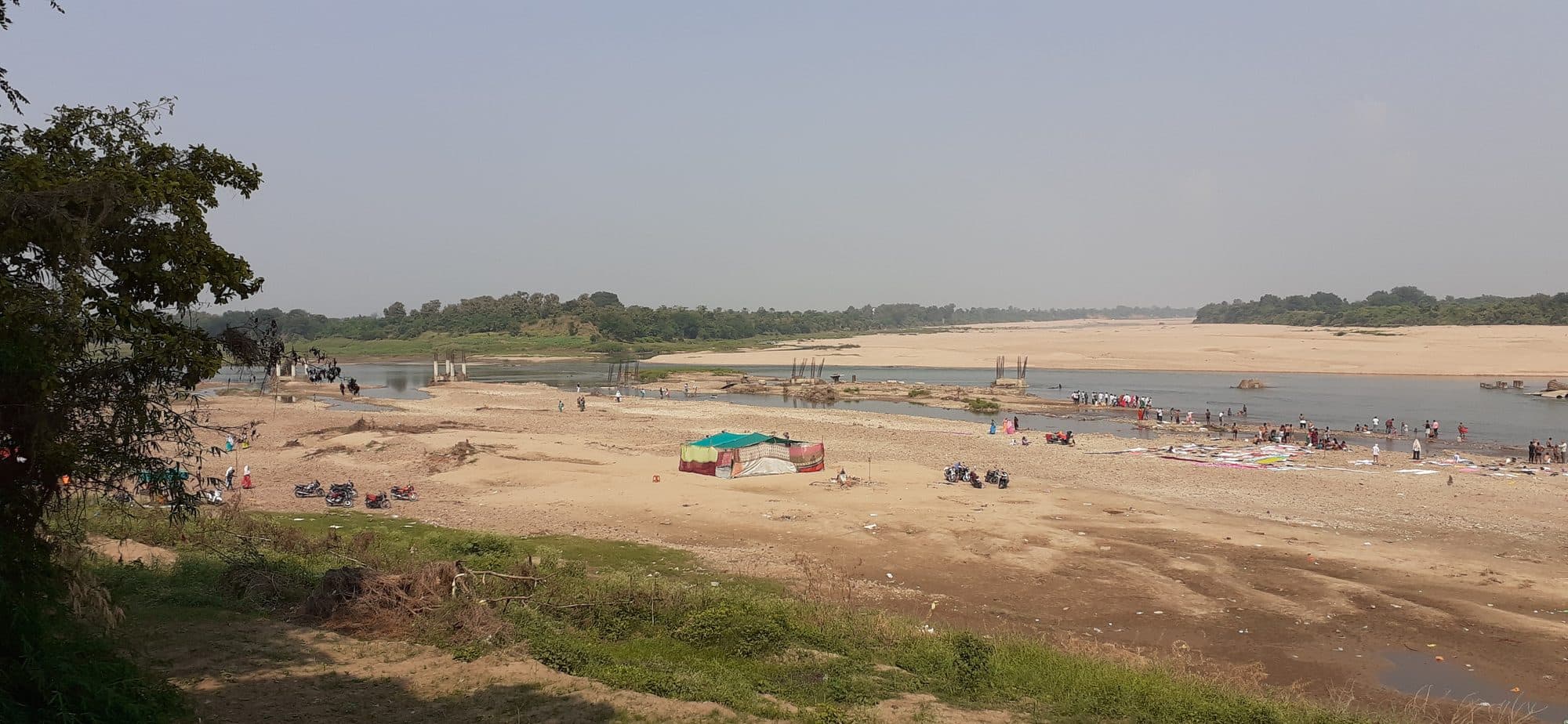 A wide river confluence in October with broad sand banks. Water runs in shallow channels on the right, crowds of people bathe and gather at the water's edge, motorbikes are parked on the sand, and a colourful tent stands in the middle of the bed. Low wooded hills line the far bank under a hazy sky.
