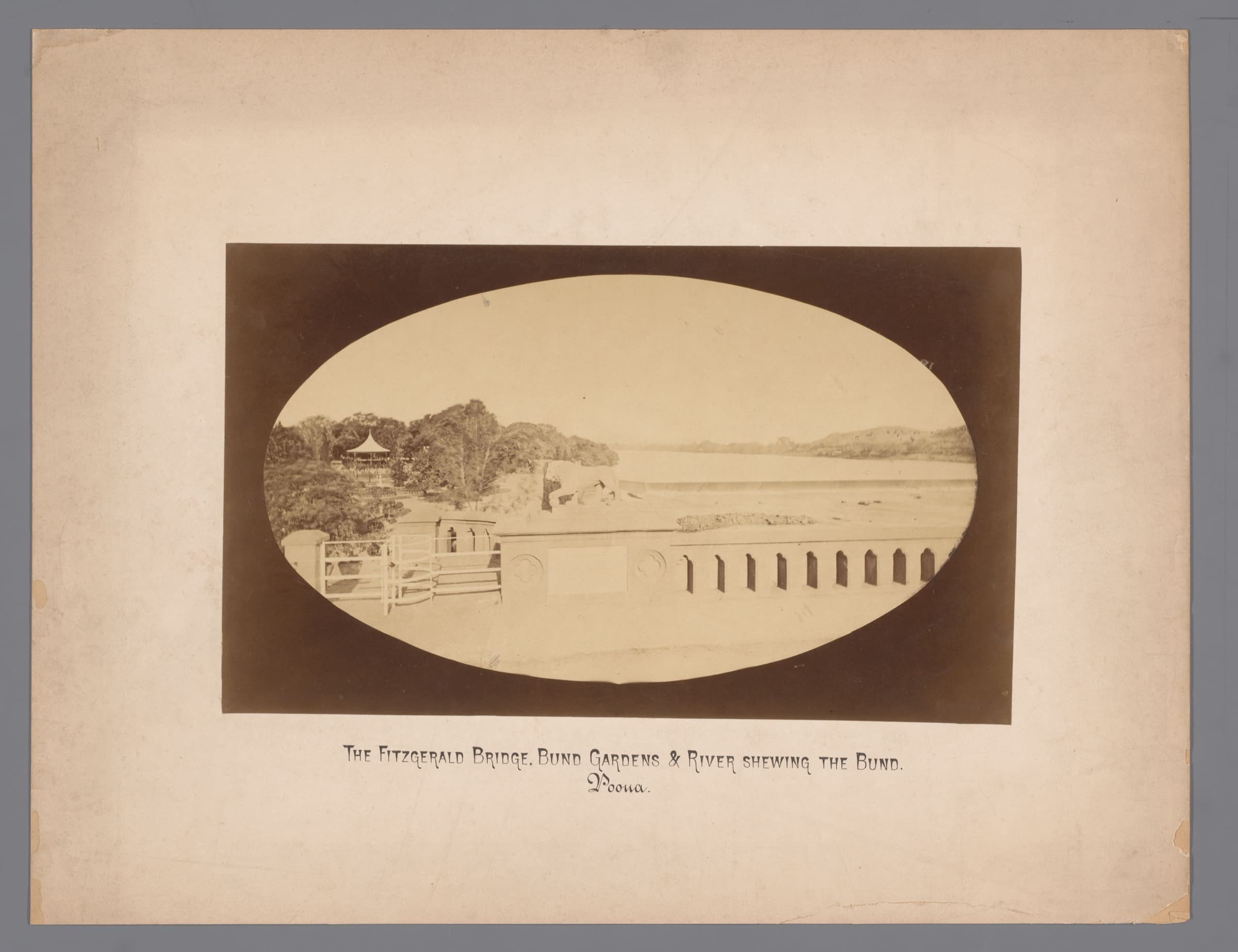 An oval-vignetted albumen print mounted on card, captioned in period lettering 'The Fitzgerald Bridge, Bund Gardens & River shewing the Bund. Poona.' A stone balustrade and the arched crest of the bund cross the foreground; behind them the Mula-Mutha widens into the pool the bund holds, with wooded banks to the left and low hills on the far shore.
