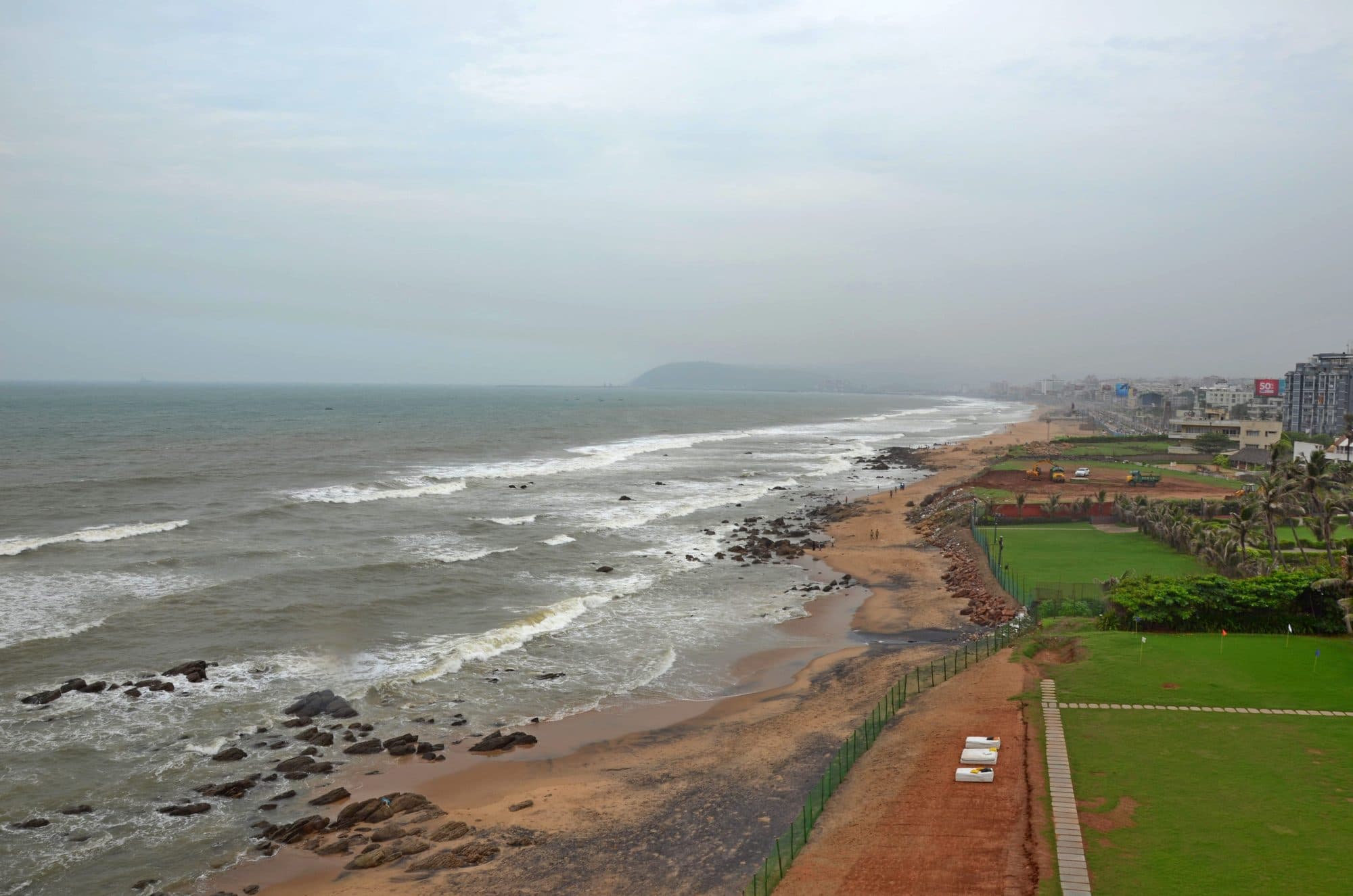 The Visakhapatnam coastline from the old Red Lighthouse in 2017: waves breaking on a rocky shore, a strip of red earth and lawns along the beach road, and a headland in the distance.