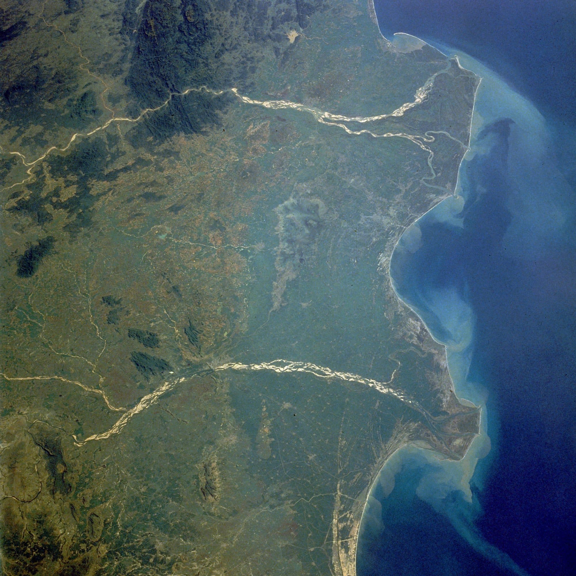 A 1989 photograph from the Space Shuttle of the Godavari and Krishna deltas: two pale river channels cross green land to a curving coast, where sediment clouds the blue Bay of Bengal.
