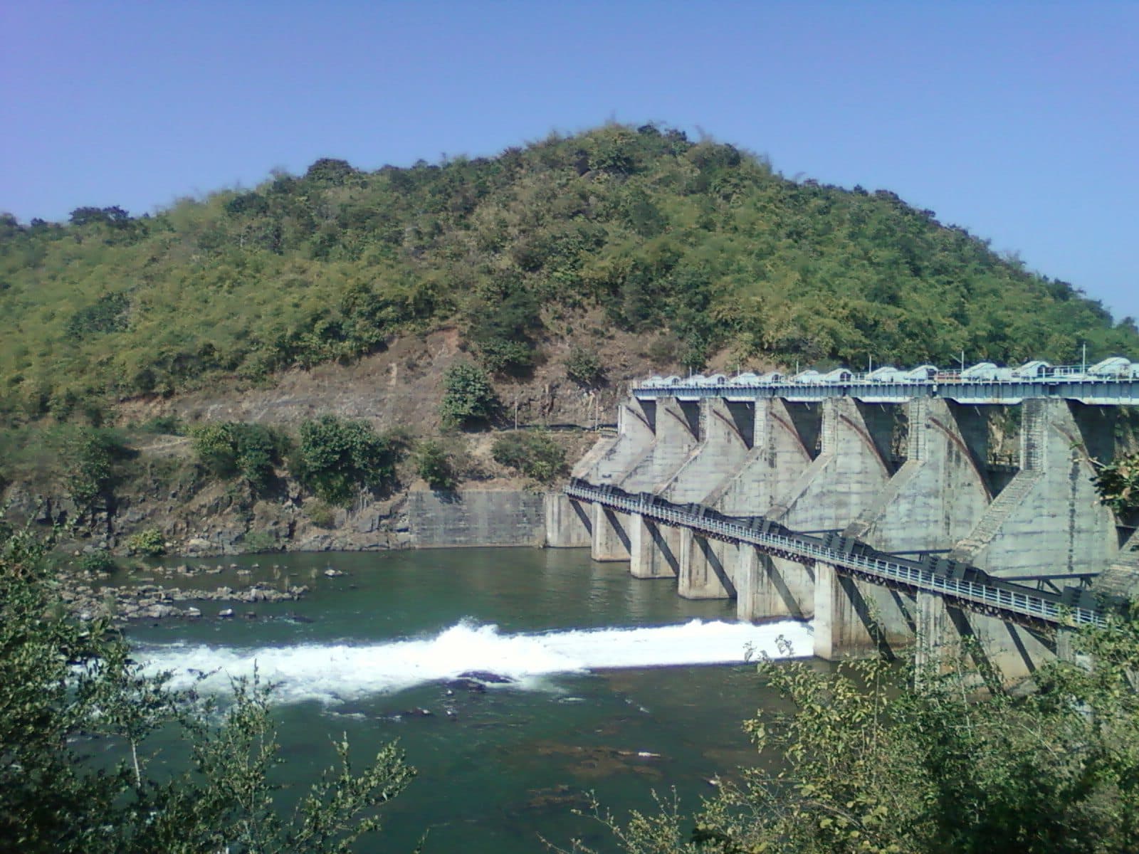 The spillway of the Yeleru dam at Yeleswaram in 2011, with water pouring through its gates below a wooded hill.